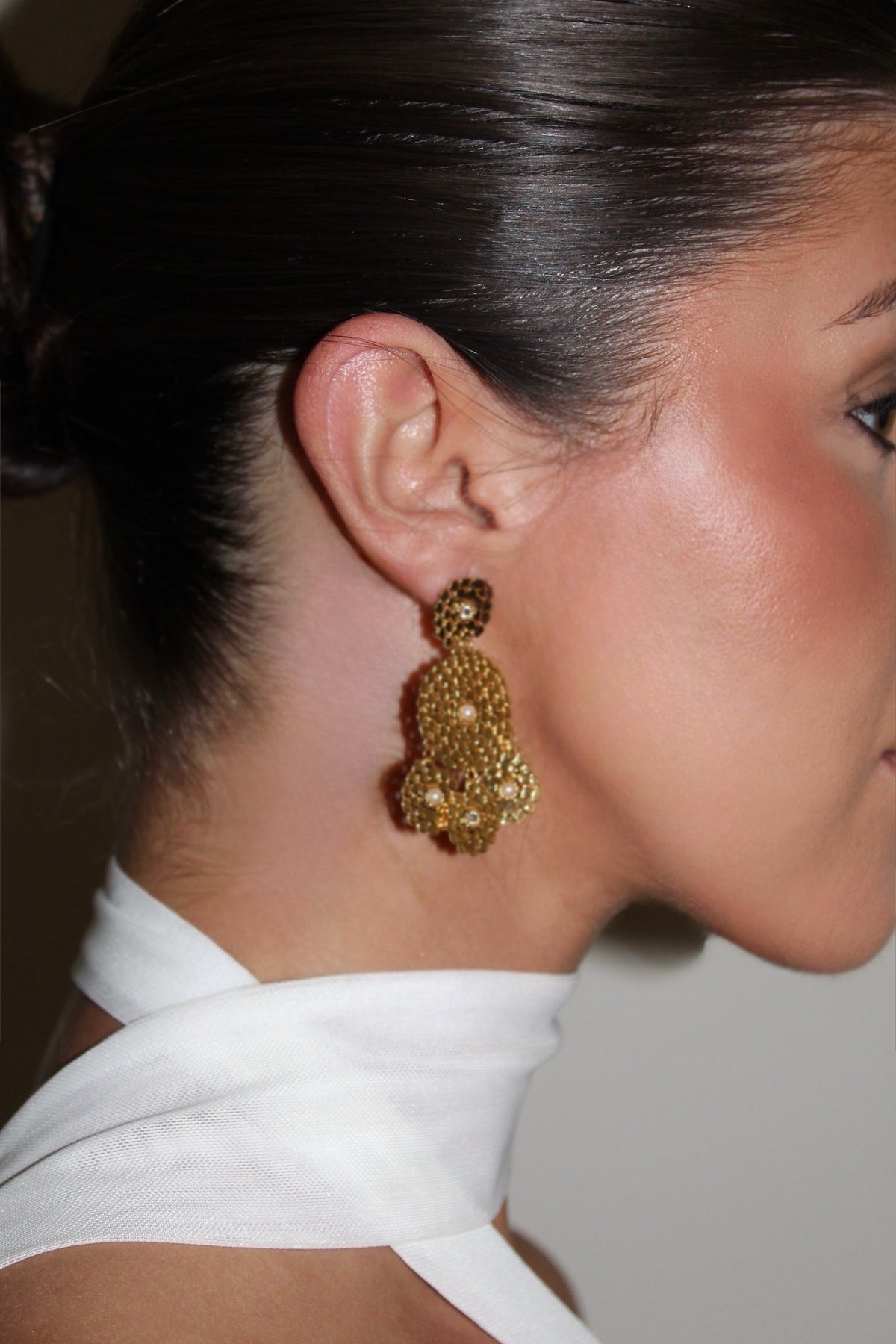 A close-up side profile of a woman wearing a large, textured gold dangle earring with small pearl-like accents. The earring hangs from her ear, showing her hair pulled back and a white collar against a neutral background.
