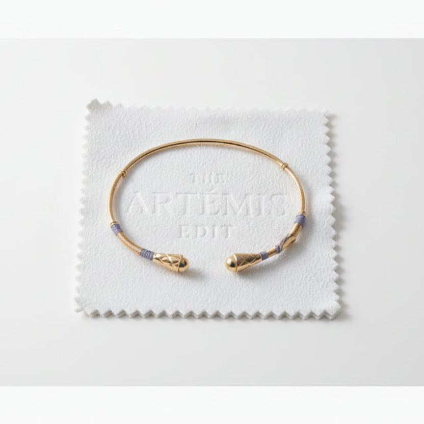 Gold bracelet with grey threads on a white cloth labeled 'The Artémis Edit' on a gray background
