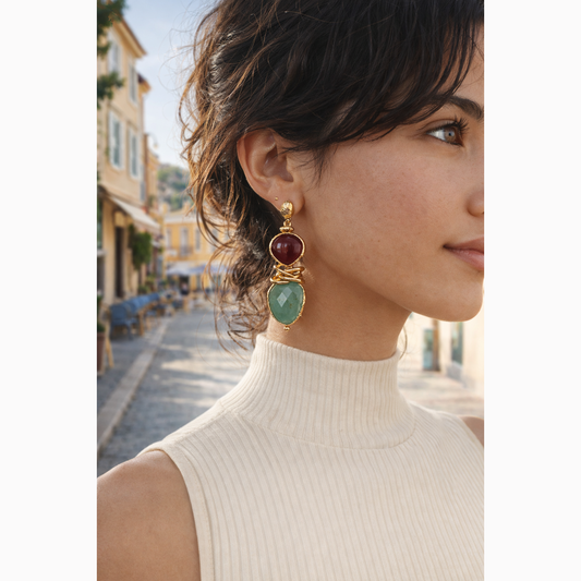 Close-up of a woman wearing gold earrings with red and green stones, outdoors.