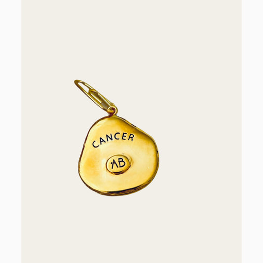 Gold pendant with 'Cancer' and 'AB' text on a light gray background