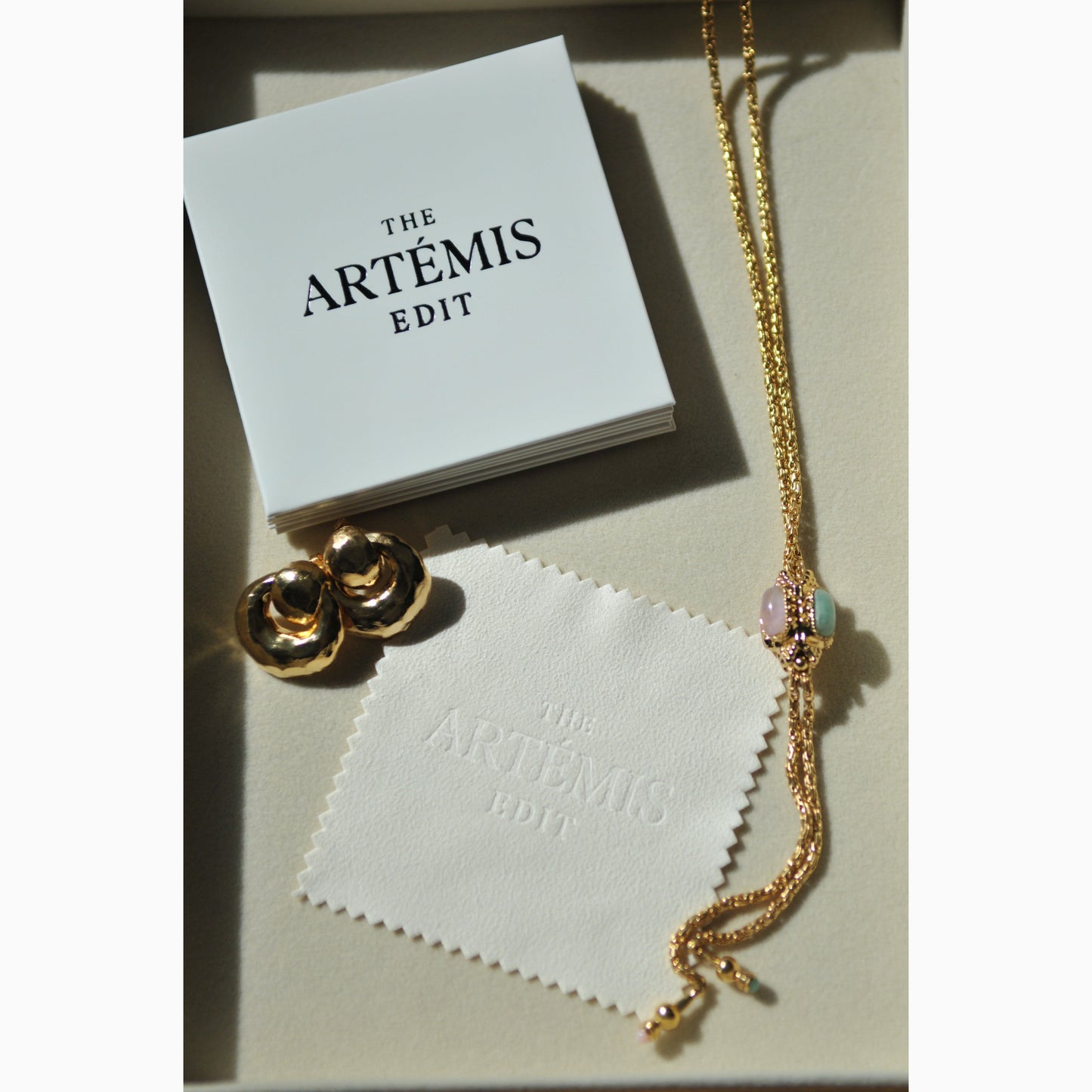 Gold necklace and earrings on a white jewellery cloth with 'The Artemis Edit' branding.