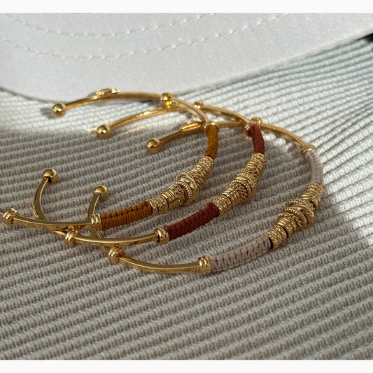 Gold and brown threaded bracelets with gold rings on a textured surface
