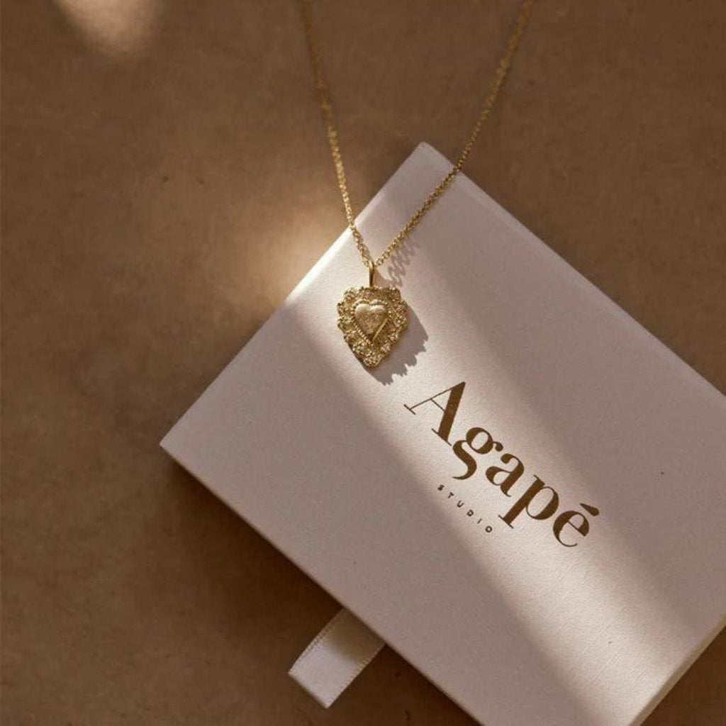 Gold necklace with a heart pendant on a 'Agapé Studio' box.