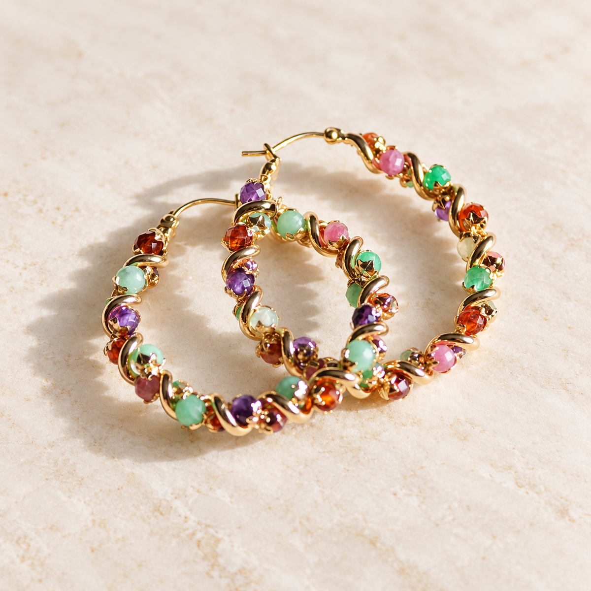 A pair of gold hoop earrings adorned with small, colorful gemstones in pink, purple, green, and orange, displayed on a light beige textured surface.