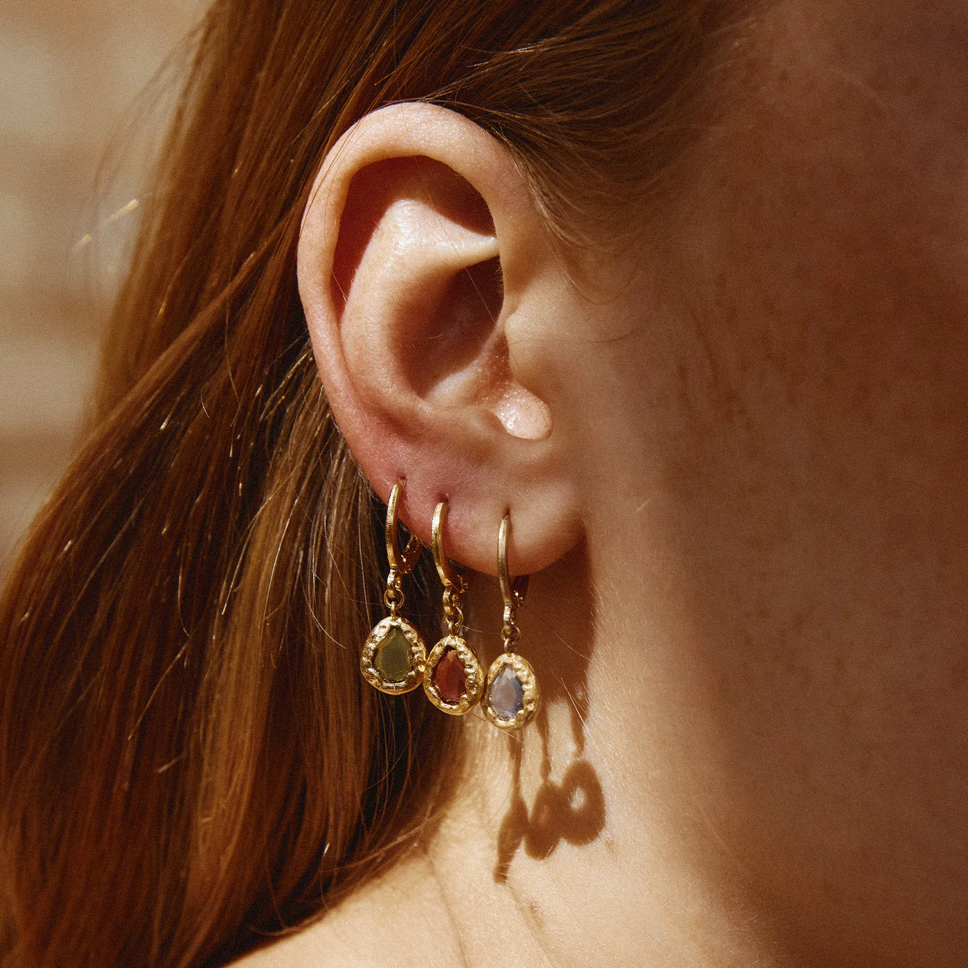 Close-up of an ear wearing gold hoop earrings with gemstones against a blurred background