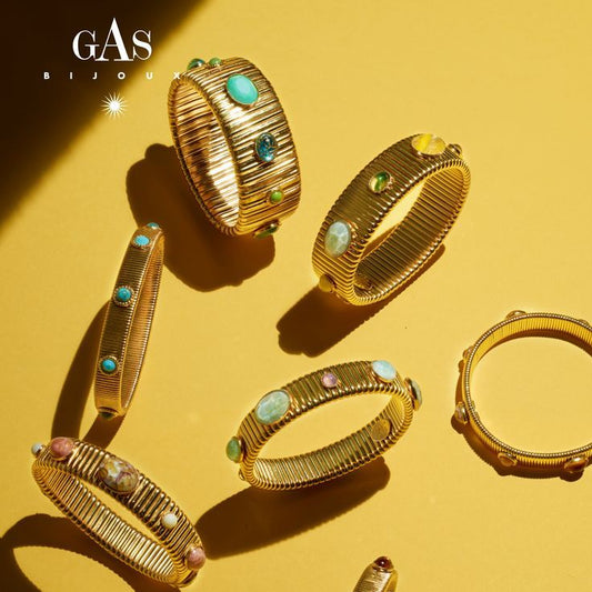 Six gold-toned bangle bracelets with textured ridges and colorful gemstone accents are arranged on a warm yellow background with dramatic shadows. The brand name GAS BIJOU is visible in the top left corner.