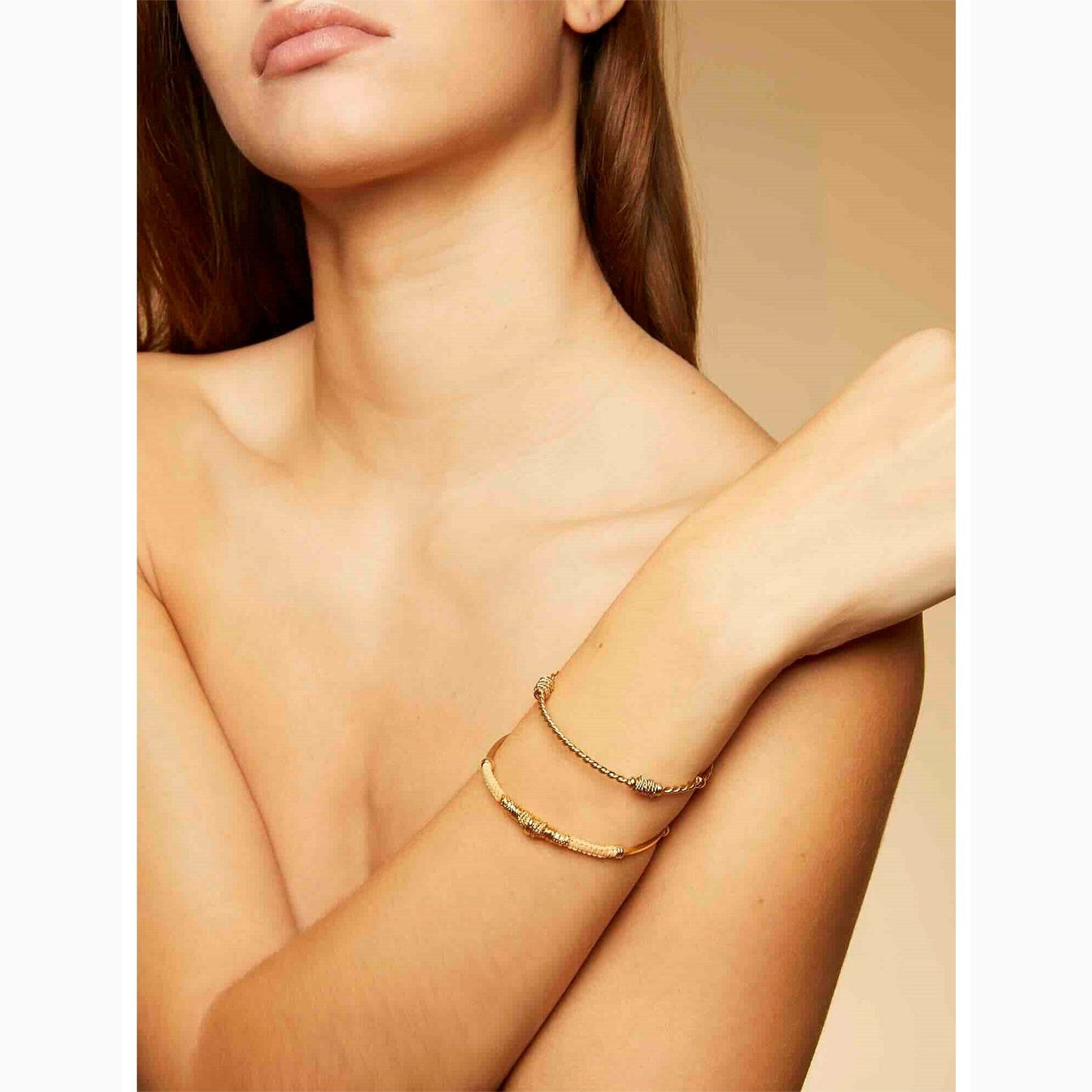 Close-up of a woman's arm wearing two gold bracelets on a beige background