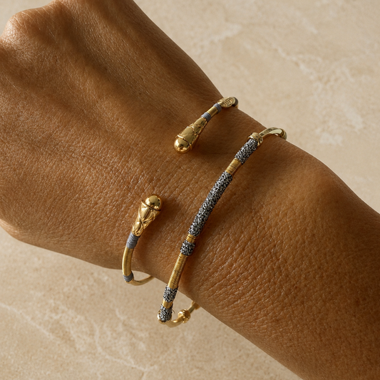 A close-up of a person's wrist wearing two gold cuff bracelets, one with small grey inlay details and the other with a textured black-and-silver pattern, set against a light beige stone background.