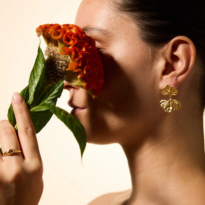 Woman holding a red flower to her face with gold earrings and ring against a beige background
