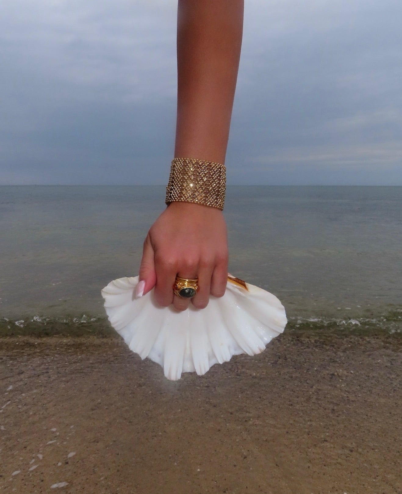 Hand holding a shell on a beach with a bracelet and ring.