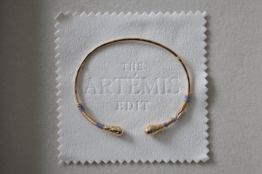 Gold bracelet with grey threads on a white cloth labeled 'The Artémis Edit' on a gray background