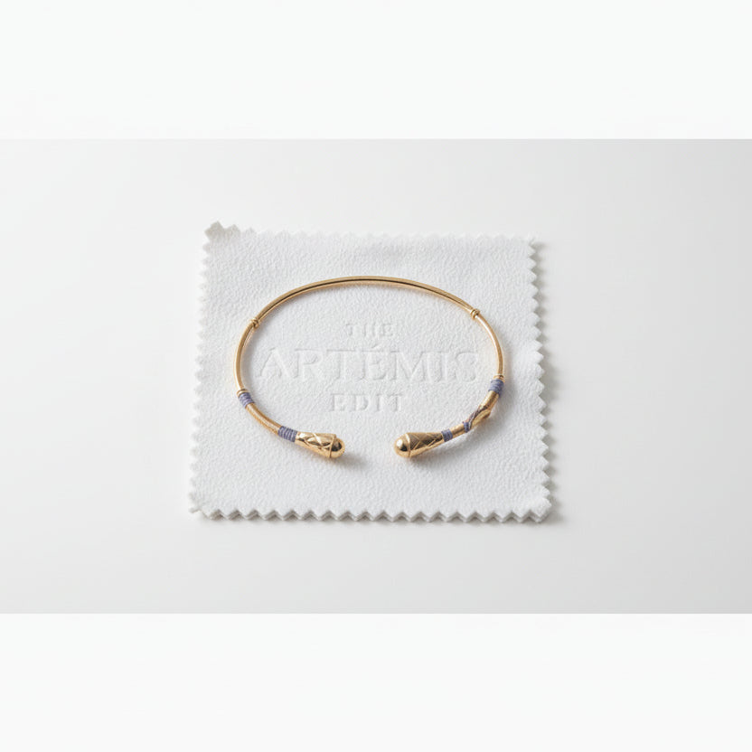 Gold bracelet with grey threads on a white cloth labeled 'The Artémis Edit' on a gray background