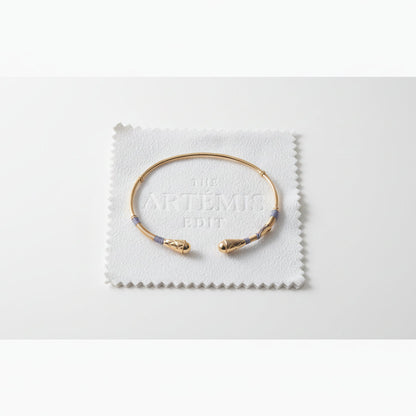 Gold bracelet with grey threads on a white cloth labeled 'The Artémis Edit' on a gray background