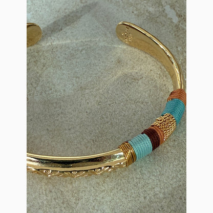 Gold bangle with colorful threads in brown and blue on a textured surface