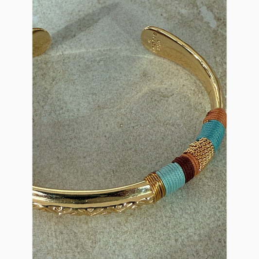 Gold bangle with colorful threads in brown and blue on a textured surface