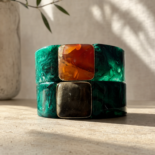 A pair of stacked resin cuff bracelets with a green and black marbled pattern, featuring square inset panels in amber-orange and dark metallic bronze, displayed on a textured beige surface with soft natural shadows.