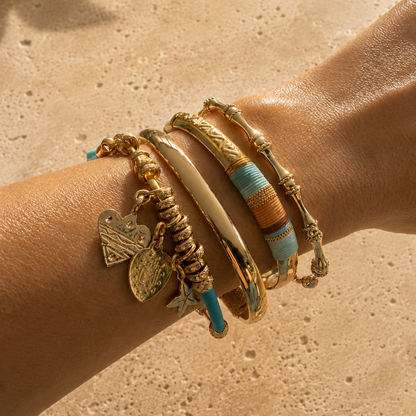 Close-up of a wrist wearing multiple gold and beaded bracelets on a beige stone surface.
