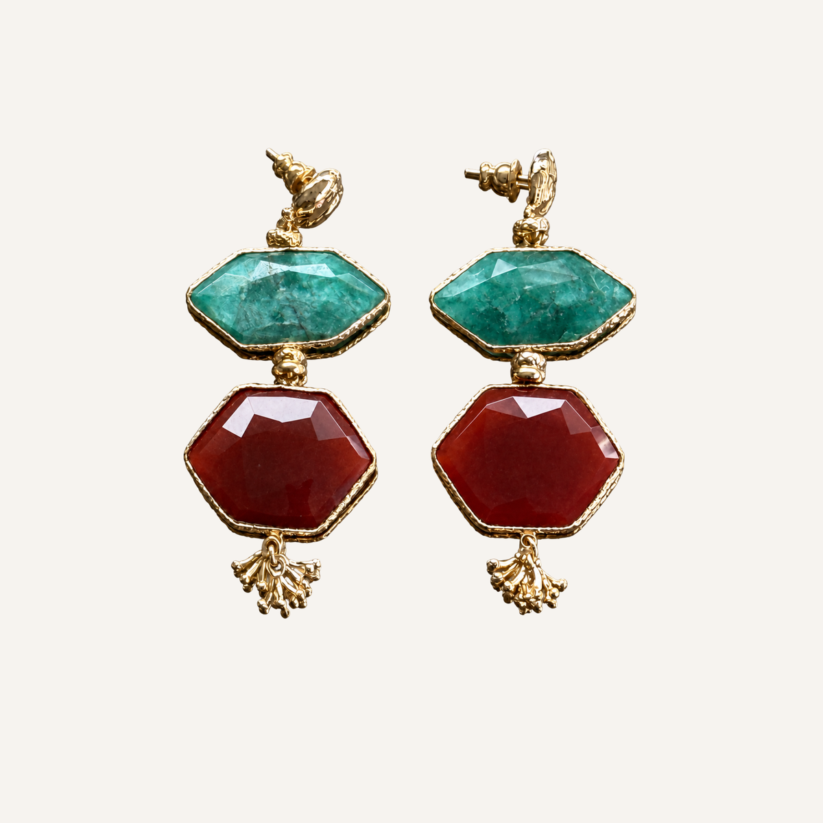 A pair of gold-tone drop earrings featuring a faceted green top stone and a larger faceted red bottom stone, displayed on a light cream background.