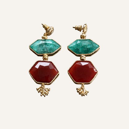 A pair of gold-tone drop earrings featuring a faceted green top stone and a larger faceted red bottom stone, displayed on a light cream background.