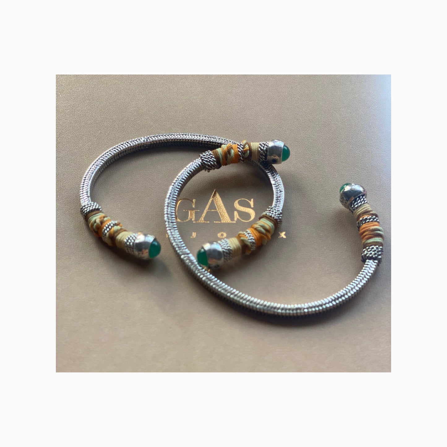 two silver bangles on a gas bijoux gift box, each have green stones at the ends and wrapped partially with coloured threads