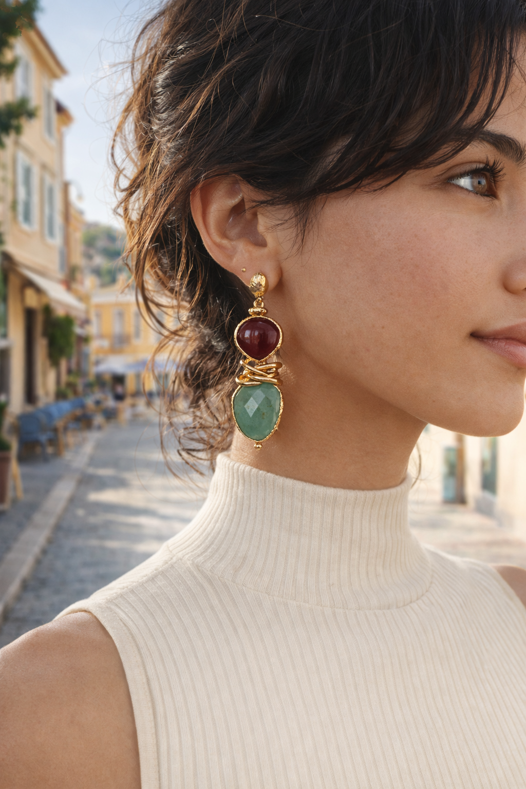 Close-up of a woman wearing gold earrings with red and green stones, outdoors.
