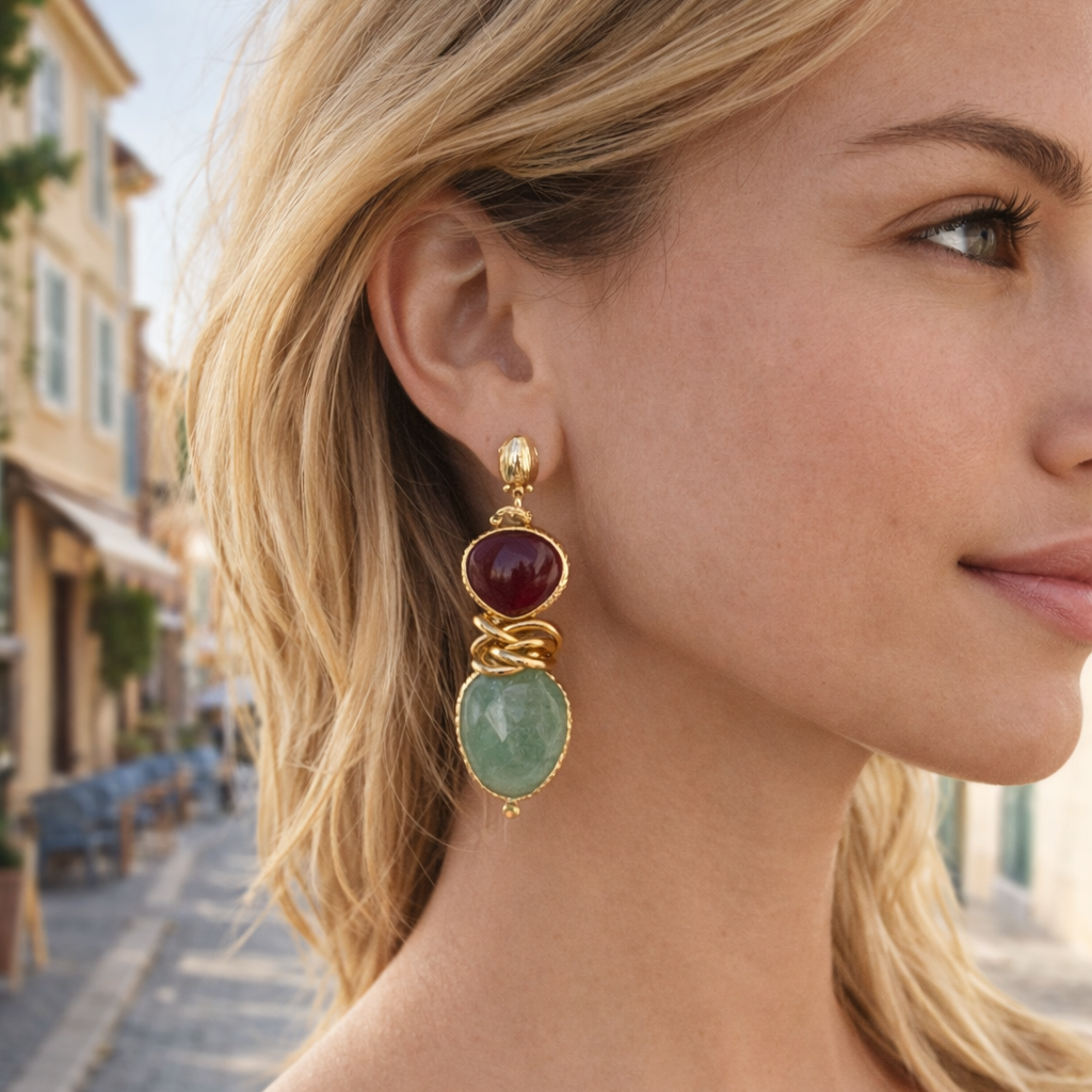 Close-up of a woman wearing gold earrings with red and green gemstones, outdoors.