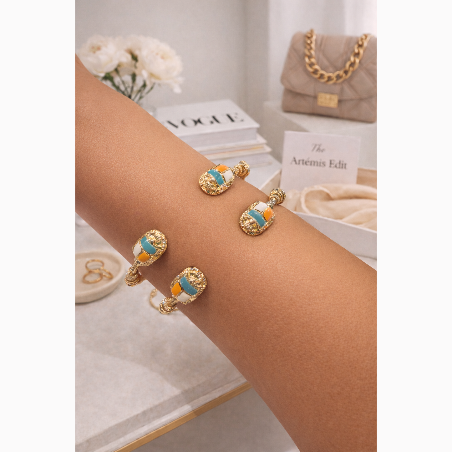 Gold bracelet with blue and orange accents on a wrist, with a blurred background of books and a handbag.