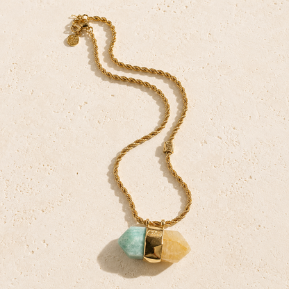 A gold chain necklace featuring a pendant with two faceted natural stones, one light aqua and one pale yellow, separated by a textured gold spacer, displayed on a light beige textured surface.