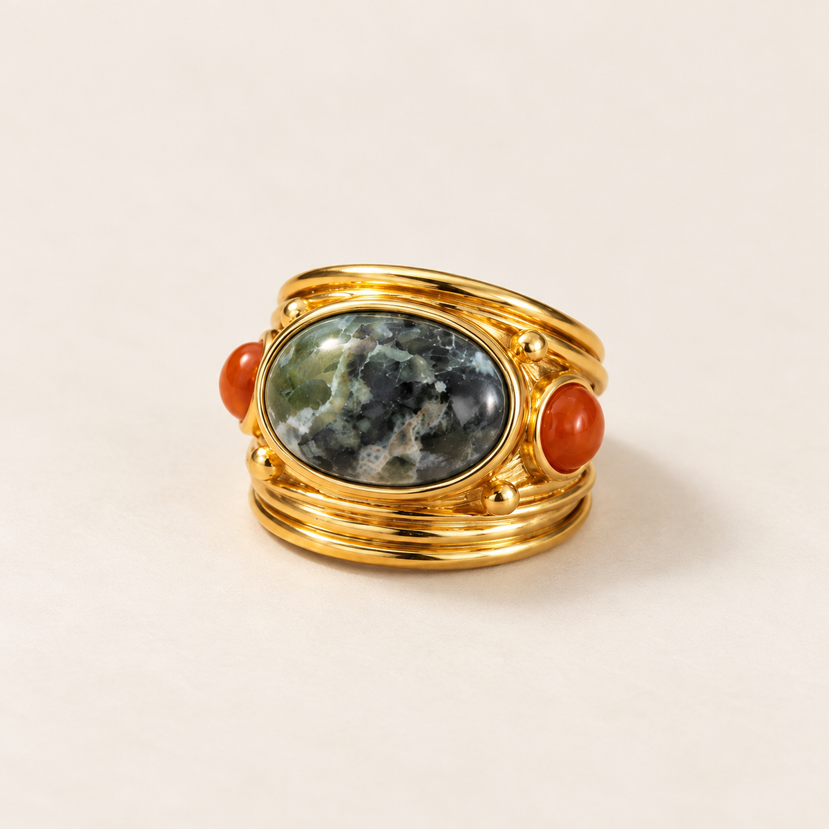 A gold ring featuring a large oval green and gray marbled stone centerpiece, flanked by two small round orange-red cabochon stones, photographed on a light beige background.