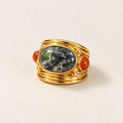 A gold ring featuring a large oval green and gray marbled stone centerpiece, flanked by two small round orange-red cabochon stones, photographed on a light beige background.