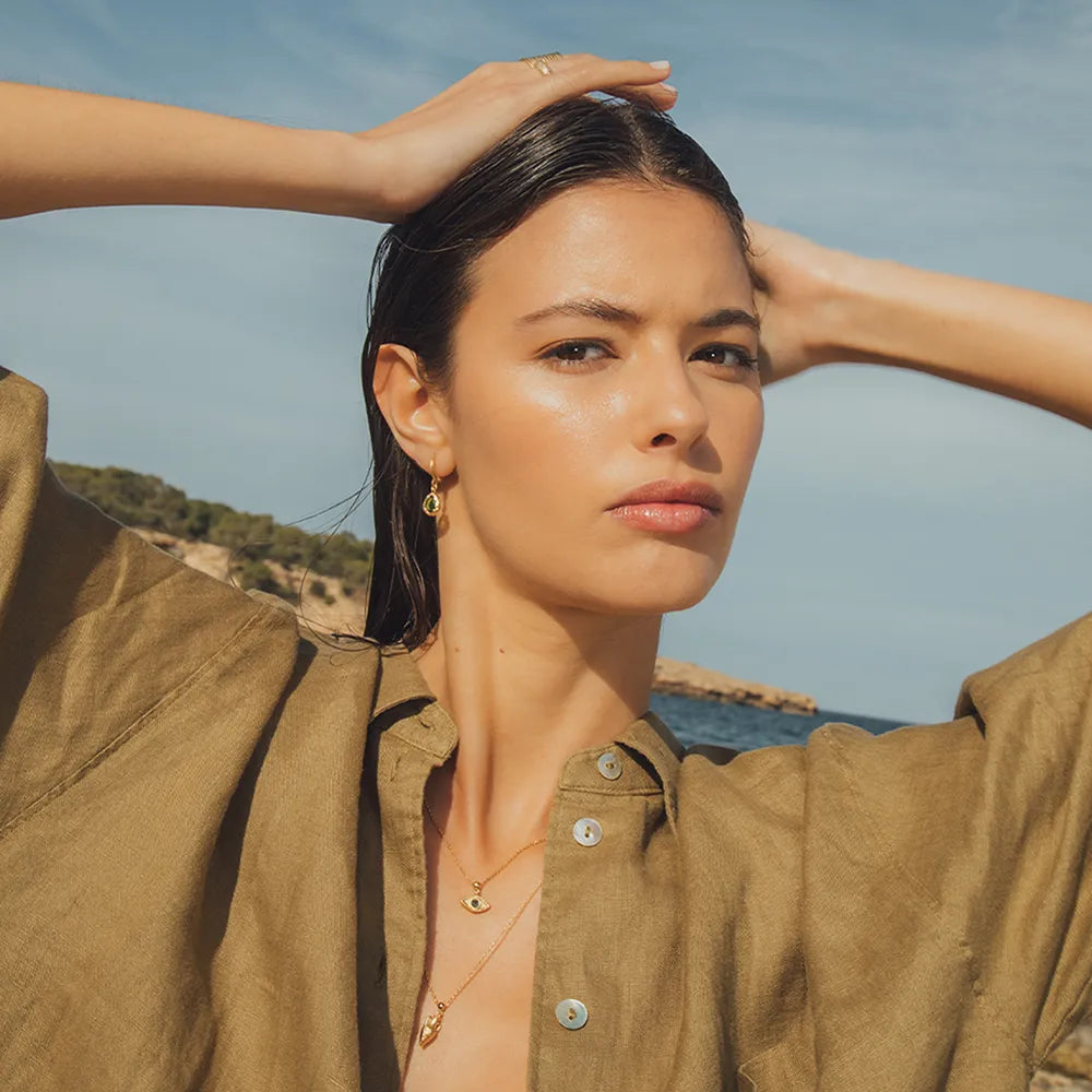 Woman in a brown shirt with hands in her hair against a blue sky wearing gold earrings with green stones at Artemis Edit