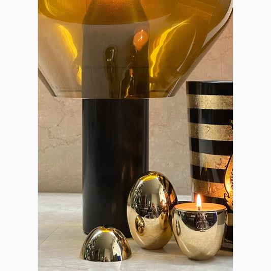 An egg-shaped gold candle with a reflective surface, placed on a marble surface, with other decorative items in the background.