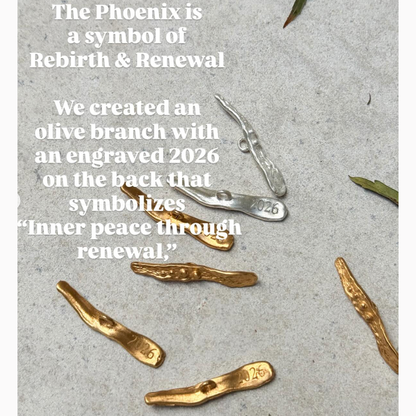 Gold and silver olive branch pendants with engraved '2026' on a textured surface.