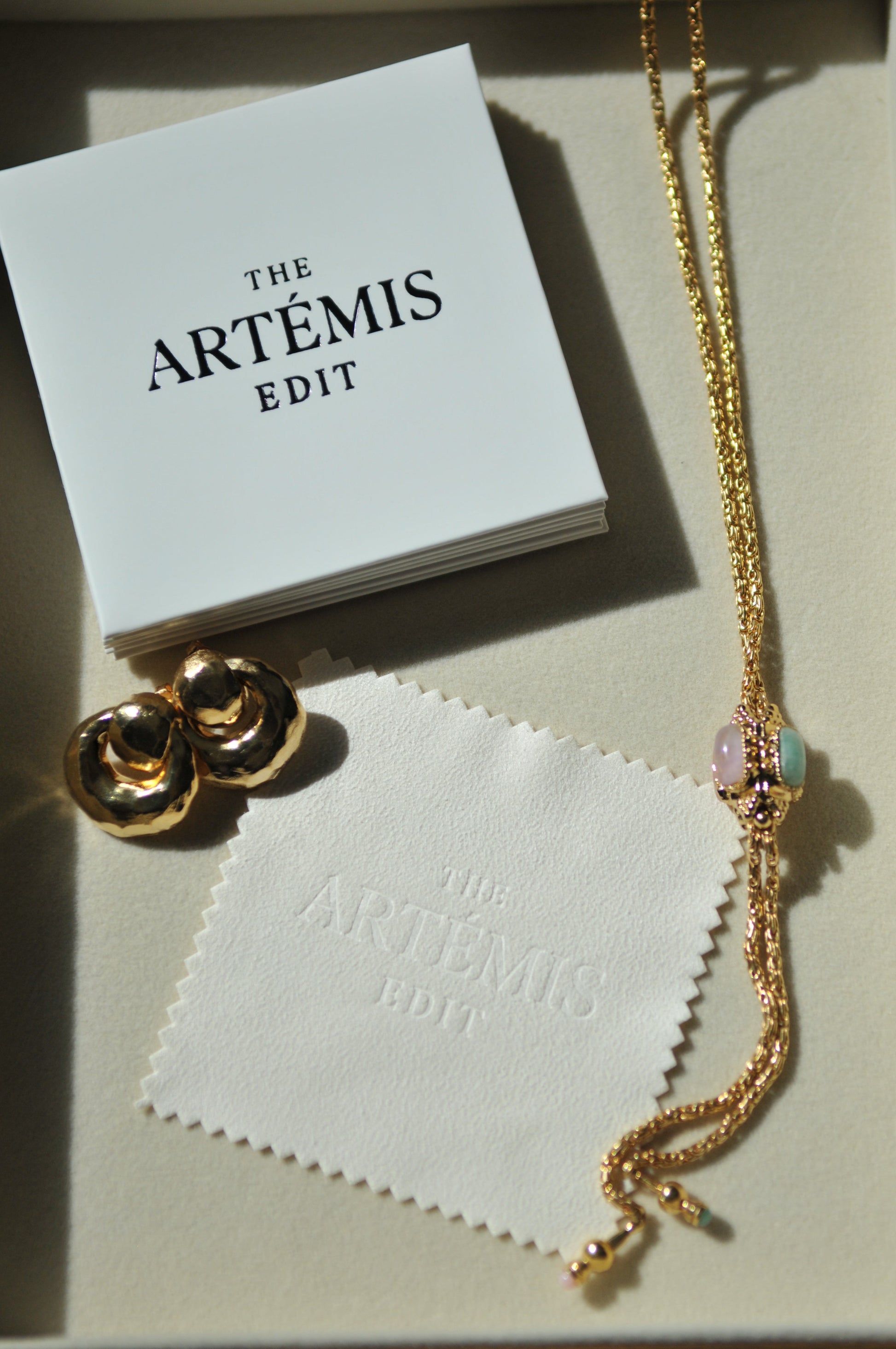Gold necklace and earrings on a white jewellery cloth with 'The Artemis Edit' branding.