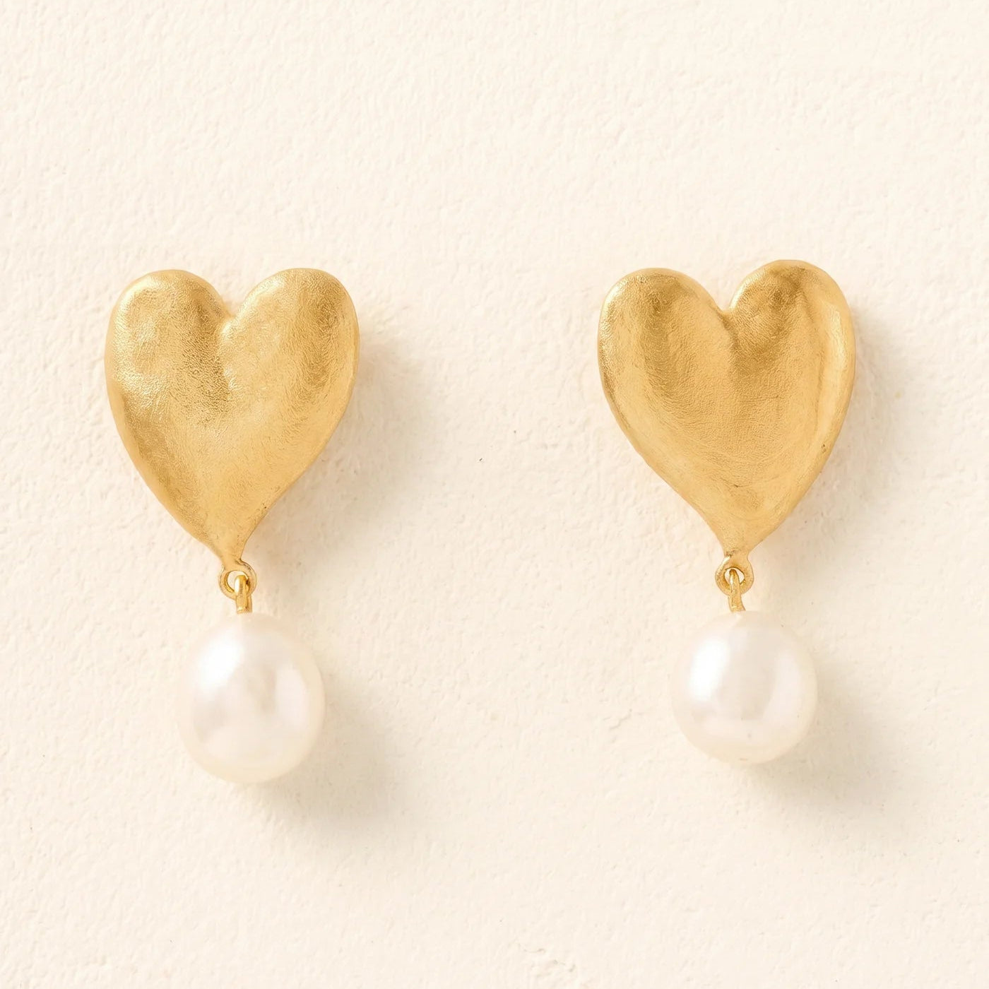 Gold heart-shaped earrings with pearl drops on a light beige background