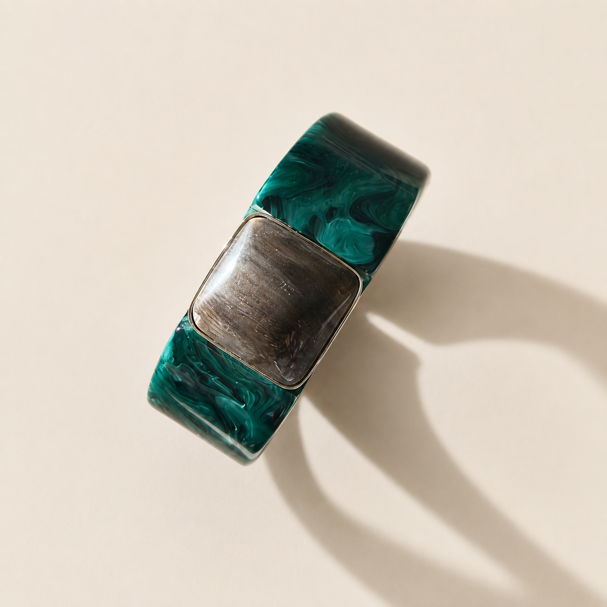 A modern bangle featuring a thick, square-shaped band with a marbled green and black resin pattern and a central square bezel-set gold platedinlay, photographed on a warm beige background with a soft shadow.