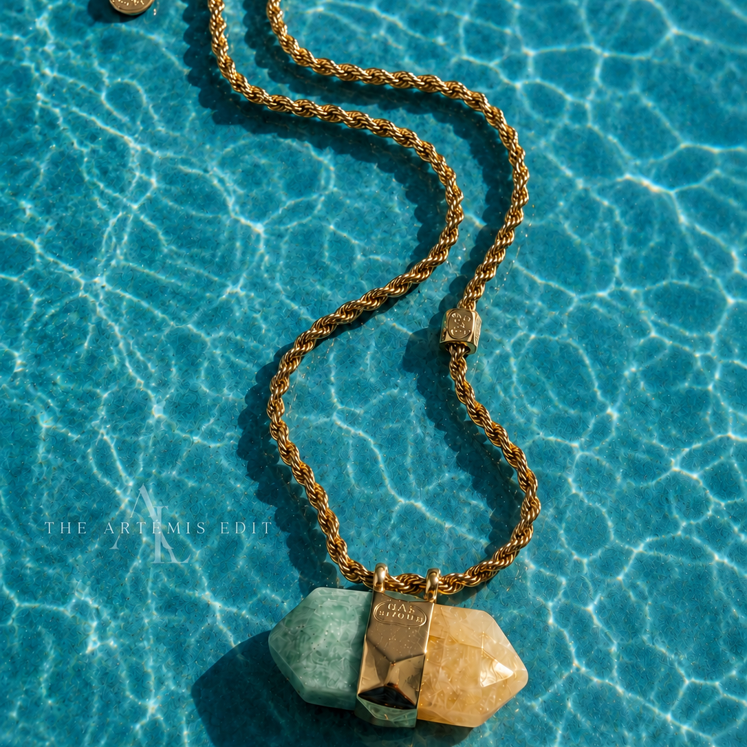 A gold chain necklace with three geometric, faceted stone pendants in green, bronze, and tan, resting on bright blue pool water with sunlight and ripples.