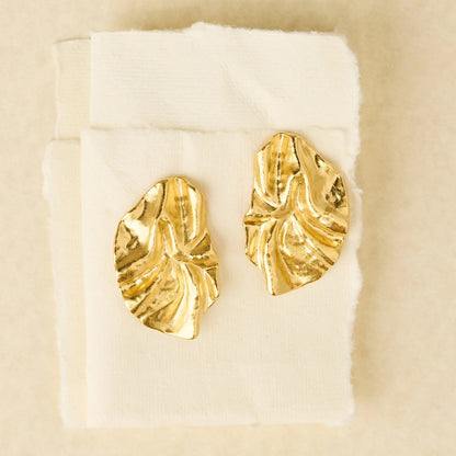 Gold leaf-shaped earrings on a white cushion with a beige background