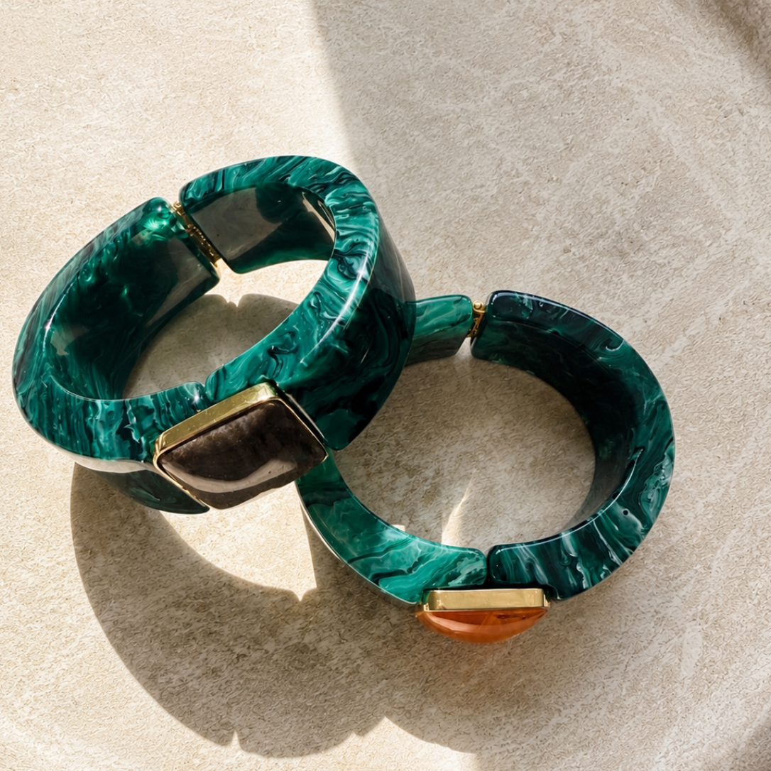 Two chunky circular bangles made of green marbled resin with gold-tone metal accents, displayed on a textured beige ceramic plate.