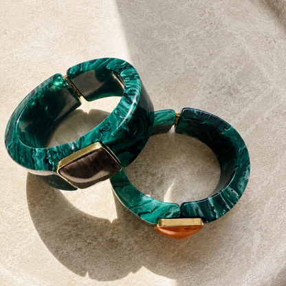 Two chunky circular bangles made of green marbled resin with gold-tone metal accents, displayed on a textured beige ceramic plate.