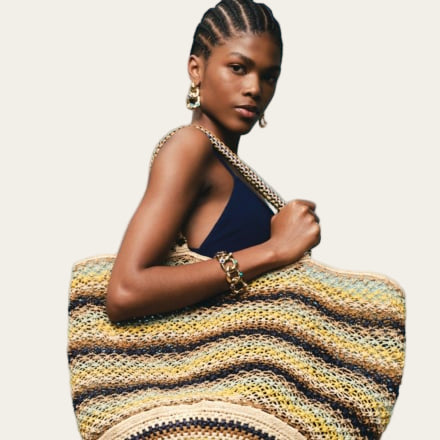 Woman holding a striped bag against a blue background wearing gold earrings and bracelet from The Artemis Edit