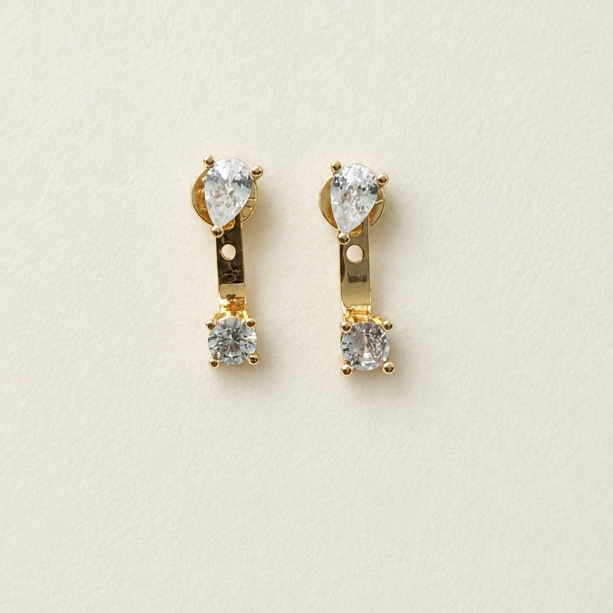 A pair of gold drop earrings with pear-cut clear crystals on the top and round clear crystals on the bottom, displayed on a textured off-white background with 'THE ARTEMIS EDIT' text at the bottom.