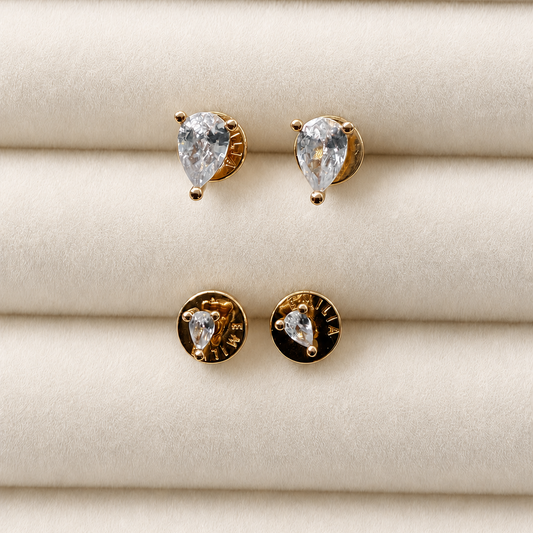 A pair of gold heart stud earrings with clear pear-cut gemstones, displayed on a cream-colored textured jewelry stand. One earring is shown from the front with the heart stone, and the other is turned to show the gold back with a small gold accent.