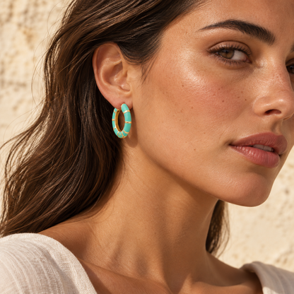 A close-up of a woman wearing a turquoise and gold striped hoop earring, set against a textured beige wall.