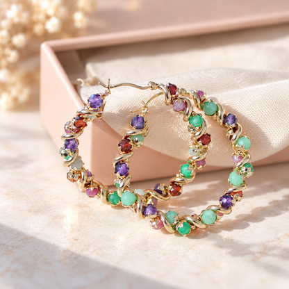 Gold hoop earrings adorned with colorful round gemstones in purple, green, red, and pink, displayed on a beige fabric surface next to a soft pink jewelry box.