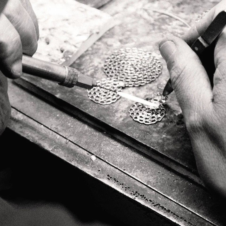 Black and white close-up of a jeweler's hands using tools to work on small circular metal pieces on a workbench.