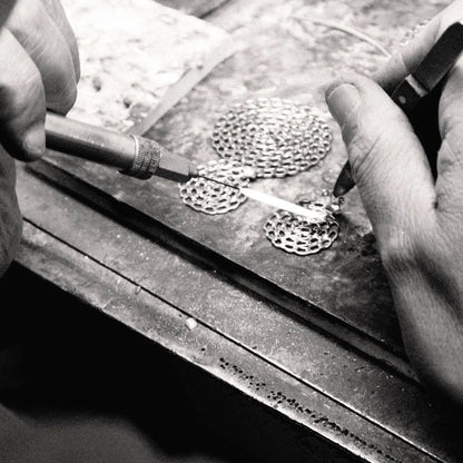 Black and white close-up of a jeweler's hands using tools to work on small circular metal pieces on a workbench.