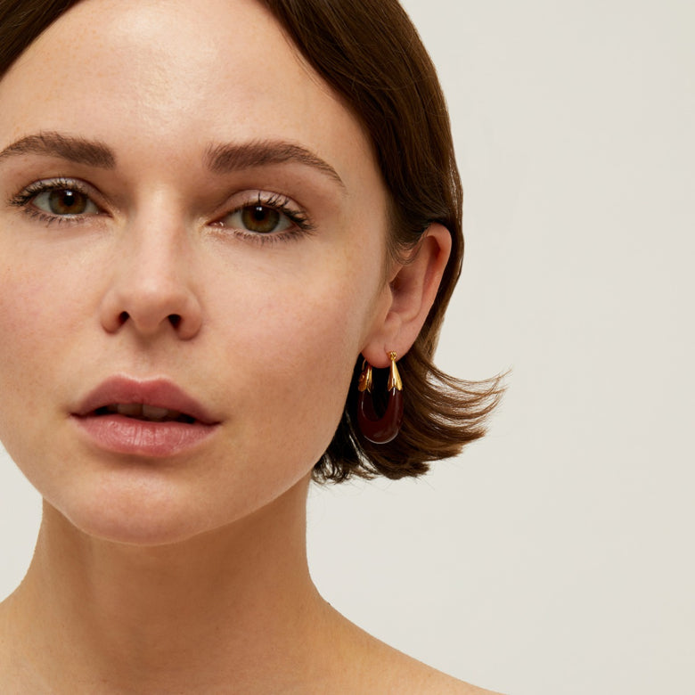 A pair of gold-tone drop earrings with a smooth finish and dark maple resin accents on a model