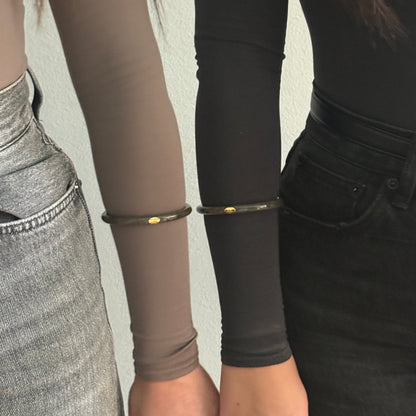 Close-up of two people standing side by side wearing slim metallic bangles on their forearms. One bangle is khaki on a taupe long-sleeve top, and the other is khaki on a black long-sleeve top.