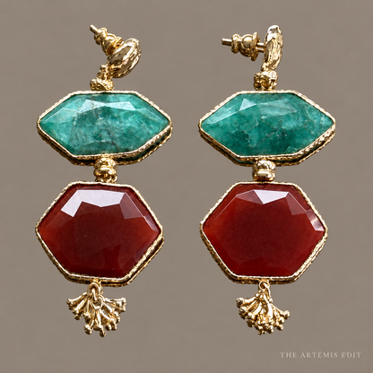 A pair of gold drop earrings featuring a faceted green top stone and a larger faceted red bottom stone, each with a small gold tassel charm, shown against a solid taupe background.